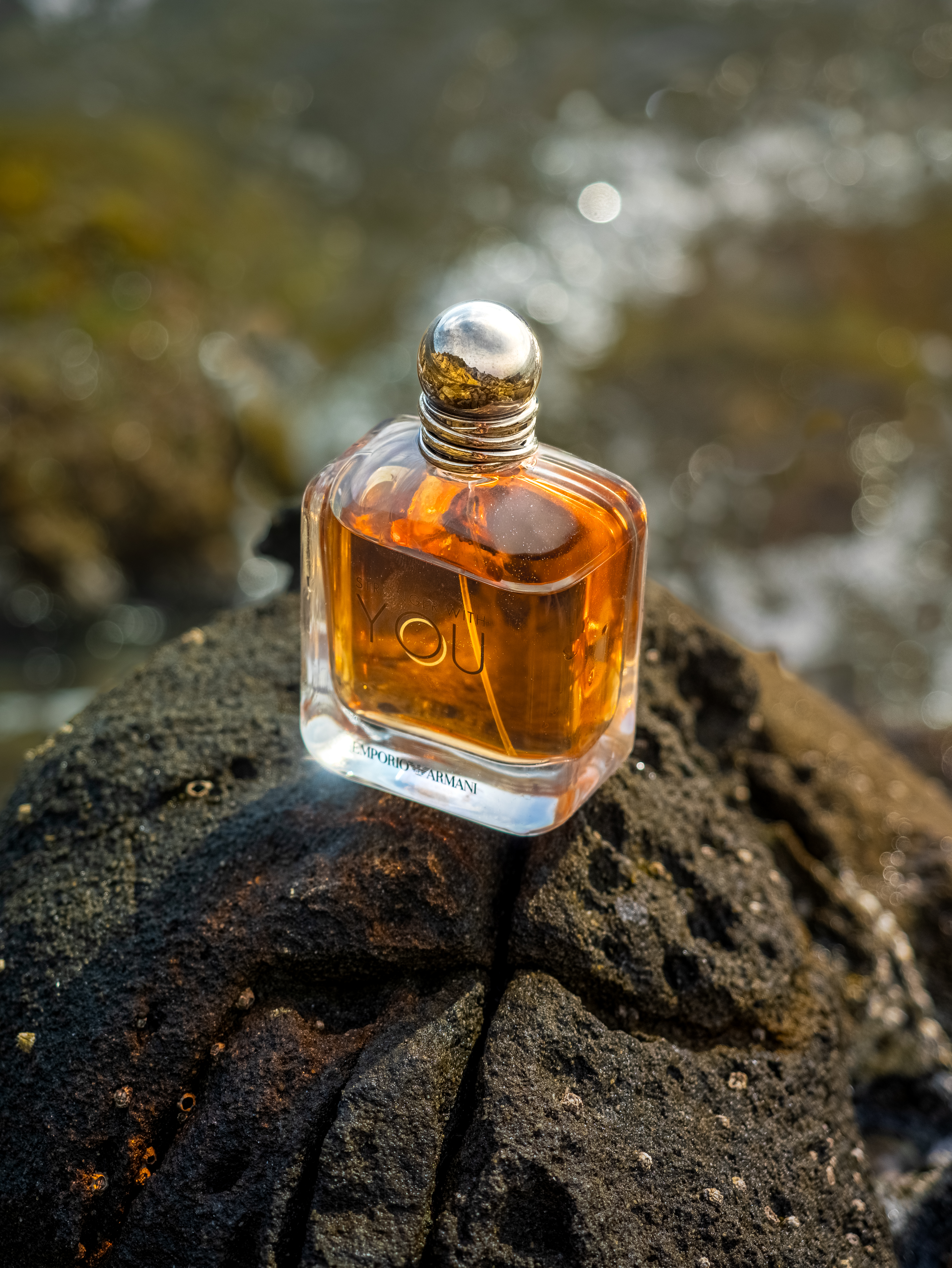 Profumo Armani Stronger With You su roccia bagnata con riflessi acqua e bokeh dorato - still life fragranza estiva mediterranea